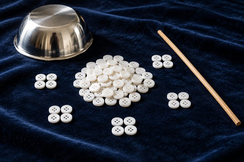 深藍色絨布上一堆白色鈕扣，旁邊幾組四顆一組的鈕扣、一個不鏽鋼碗和一根竹籤
